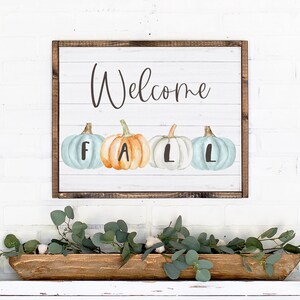 May include: A rectangular wall sign with a white shiplap background and a dark wood frame. The sign features the word Welcome in black script above four watercolour pumpkins in shades of blue and orange, each marked with the letters F, A, L, L.