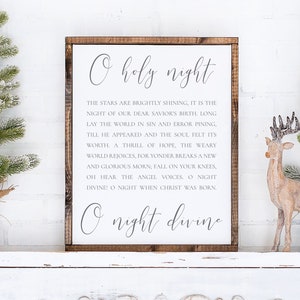 以下が含まれることがあります： 白い木製の看板に、クリスマスキャロル「O Holy Night」の歌詞が黒で印刷されています。看板は白い暖炉の上に置かれ、小さな雪が積もった人工のクリスマスツリー2本と木製のトナカイの置物が置かれています。