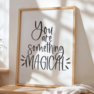 Puede incluir: Un cuadro enmarcado con las palabras "You are something MAGICAL" en escritura negra. La impresión está en un marco de madera clara y descansa sobre una cómoda de madera. Una manta blanca está sobre la cómoda.