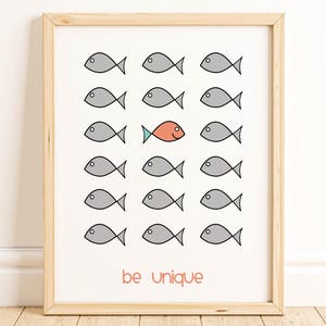 以下が含まれることがあります： グレーの魚の群れとオレンジ色の魚が描かれた額入りのアートプリント。「be unique」というフレーズが魚の下に赤で書かれています。アートワークは明るい木製のフレームに入っています。