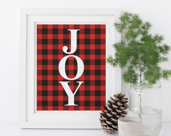 May include: A framed print with the word "JOY" in white letters against a red and black buffalo plaid background. A glass jar with greenery, a pine cone, and a small concrete candle holder are next to the frame. The frame is white.
