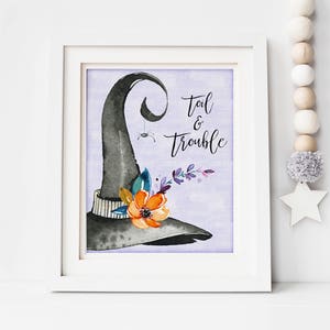 May include: A framed watercolor print featuring a black witch's hat with an orange flower and the words "Toil & Trouble." The artwork has a lavender background and is in a white frame. A decorative garland hangs nearby.