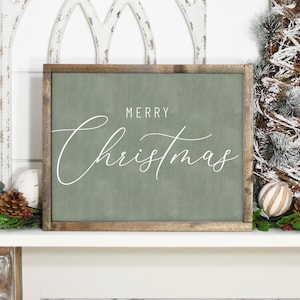 Pode incluir: Uma placa de madeira verde com o texto "Merry Christmas" em escrita cursiva branca. A placa est&aacute; sentada em uma lareira branca com uma pequena &aacute;rvore de pinho nevada e outras decora&ccedil;&otilde;es de Natal.