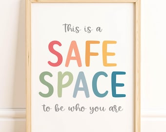 May include: A framed print with the words "This is a SAFE SPACE to be who you are" in colorful letters. The frame is light wood, and the print is displayed against a white wall and wooden floor.