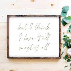 Puede incluir: Un letrero enmarcado de madera con el texto "but I think I love Fall most of all" en cursiva. Decoraciones otoñales, calabazas y hojas de eucalipto sobre una superficie de madera clara.