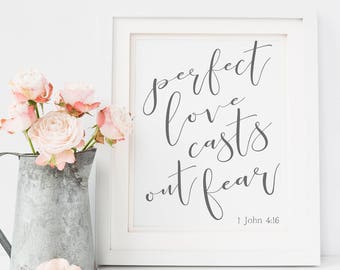 May include: A white framed print with the text "perfect love casts out fear" and the scripture reference "1 John 4:18". The print is sitting on a white surface with a metal pitcher filled with pink flowers.