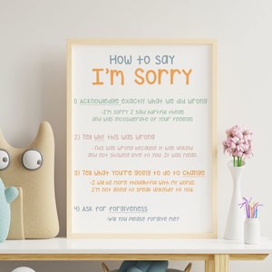 Puede incluir: Un póster enmarcado con el título "Cómo decir Lo siento" con cuatro pasos enumerados en texto naranja y azul. El póster está en un estante blanco con un jarrón de flores rosas, una cesta de juguetes de peluche y una pila de libros.