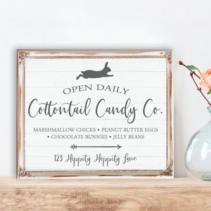 May include: A white sign with a distressed wood look, featuring a silhouette of a bunny rabbit hopping. The sign reads "OPEN DAILY Cottontail Candy Co. MARSHMALLOW CHICKS PEANUT BUTTER EGGS &bull; CHOCOLATE BUNNIES JELLY BEANS 123 Hippity Hoppity Lane"