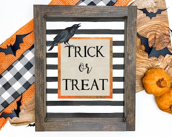 Puede incluir: Un letrero de Halloween enmarcado con las palabras "Trick or Treat" y una ilustración de un cuervo negro. El letrero tiene un fondo a rayas blancas y negras y un marco marrón oscuro. Elementos decorativos: cinta naranja, murciélagos de papel y calabazas.