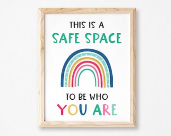 May include: A framed print with the text "THIS IS A SAFE SPACE TO BE WHO YOU ARE" above a colorful rainbow illustration. The rainbow features blue, yellow, red, and green arches. The frame is made of light-colored wood.