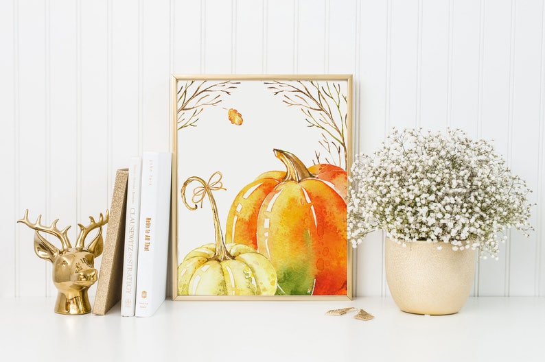 May include: A framed watercolor print featuring pumpkins in shades of orange, yellow, and green. The artwork is set against a white background and framed in gold. A gold deer head figurine, books, and a vase of white flowers are also present.