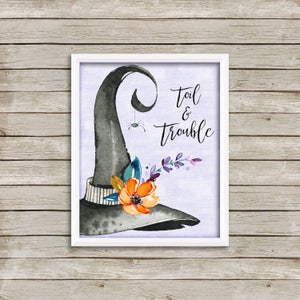 May include: A framed watercolor print featuring a black witch's hat with a spider hanging from it. The hat is adorned with orange and purple flowers. The words "Toil & Trouble" are written in elegant script.