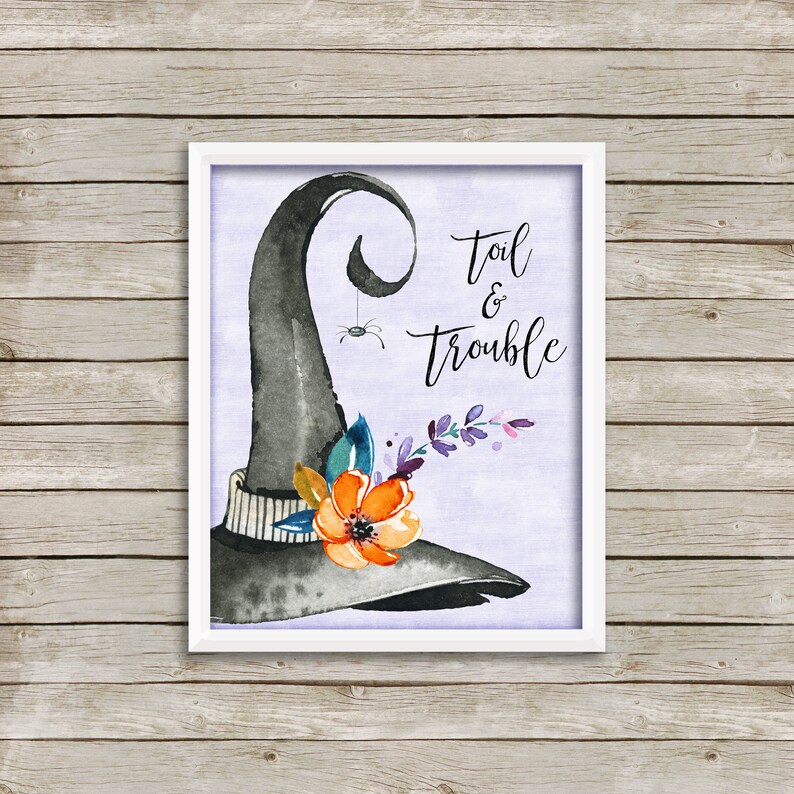 May include: A framed watercolor print featuring a black witch's hat with a spider hanging from it. The hat is adorned with orange and purple flowers. The words "Toil & Trouble" are written in elegant script.