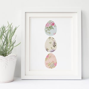 May include: A white frame with a white mat surrounding a print of three pastel-colored eggs with floral patterns. The eggs are arranged vertically in a row.