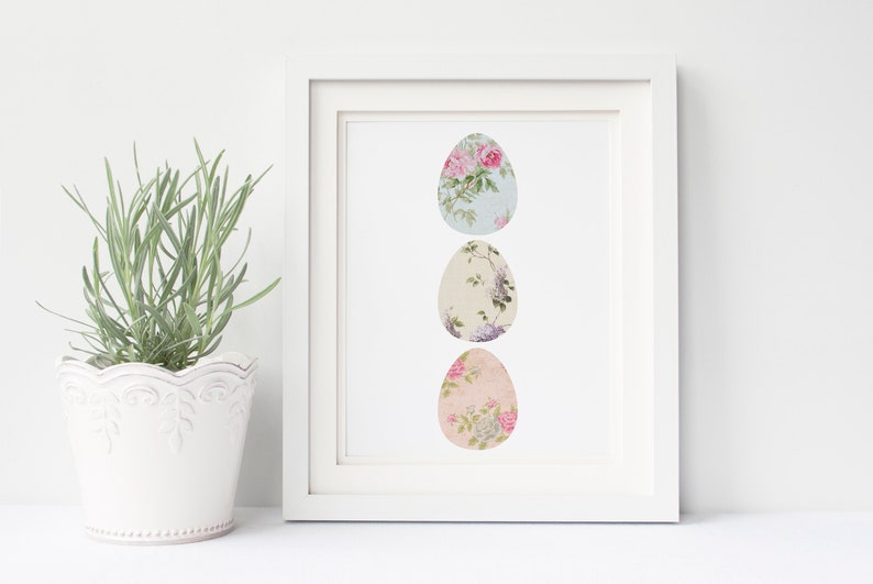 May include: A white frame with a white mat surrounding a print of three pastel-colored eggs with floral patterns. The eggs are arranged vertically in a row.