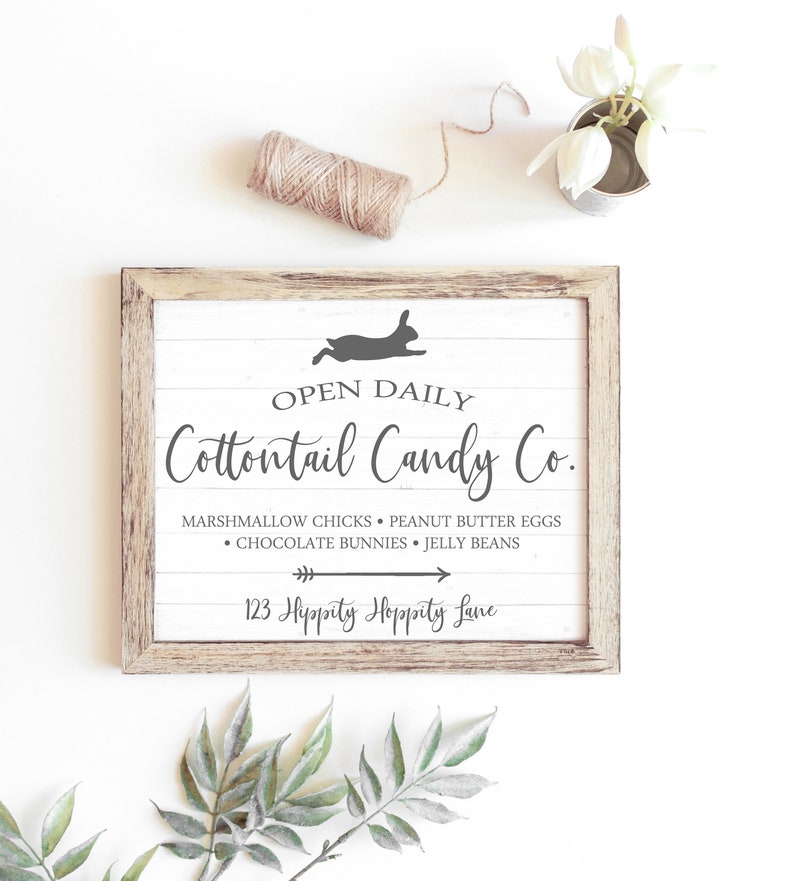May include: A white wooden sign with a brown frame, featuring the text "OPEN DAILY Cottontail Candy Co." and a silhouette of a rabbit. The sign lists the candy shop's offerings: "Marshmallow Chicks, Peanut Butter Eggs, Chocolate Bunnies, Jelly Beans." The address is listed as "123 Hippity Hoppity Lane."