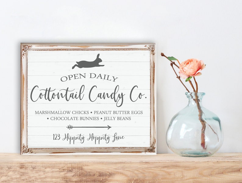 May include: A white sign with a distressed wood look, featuring a silhouette of a bunny rabbit hopping. The sign reads "OPEN DAILY Cottontail Candy Co. MARSHMALLOW CHICKS PEANUT BUTTER EGGS &bull; CHOCOLATE BUNNIES JELLY BEANS 123 Hippity Hoppity Lane"