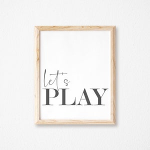 May include: A framed print with the text "let's PLAY" in black and white on a white background. The frame is made of light wood.