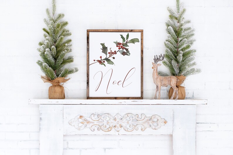 May include: A framed sign with the word "Noel" in elegant script, featuring a watercolor holly branch with red berries. Two small artificial Christmas trees and a wooden reindeer are on a white mantel.