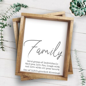 以下が含まれることがあります： 木製のフレームに入ったサインで、エレガントな筆記体で「Family」という言葉が書かれています。家族の定義が単語の下に印刷されています。フレームは明るい茶色です。サインは白い木の表面にあります。多肉植物とユーカリの小枝が背景にあります。