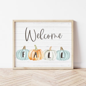 May include: A rectangular framed sign with a white wood-plank background featuring the word Welcome in black script. Below the text are four watercolour-style pumpkins in light blue, orange, and white, each marked with the letters F, A, L, L.