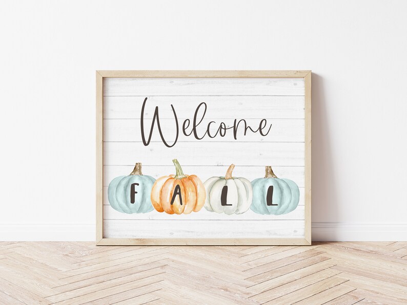 May include: A rectangular framed sign with a white wood-plank background featuring the word Welcome in black script. Below the text are four watercolour-style pumpkins in light blue, orange, and white, each marked with the letters F, A, L, L.