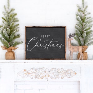 Pode incluir: Uma placa preta enquadrada com o texto "Merry Christmas" em escrita branca sobre fundo preto. A placa est&aacute; sentada em uma lareira branca com duas pequenas &aacute;rvores de Natal artificiais cobertas de neve e uma figura de rena de madeira.