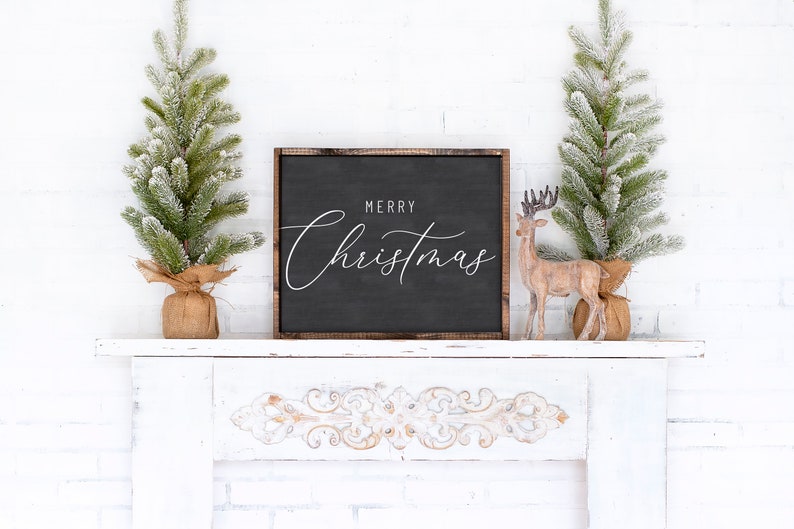 Pode incluir: Uma placa preta enquadrada com o texto "Merry Christmas" em escrita branca sobre fundo preto. A placa est&aacute; sentada em uma lareira branca com duas pequenas &aacute;rvores de Natal artificiais cobertas de neve e uma figura de rena de madeira.