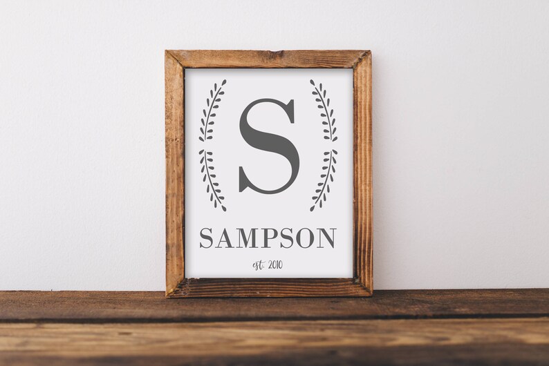May include: A framed wooden sign with a white background features a large gray letter "S" and the name "SAMPSON" in a serif font. Decorative leaf branches flank the letter. The text "est. 2010" is below the name. The frame is made of dark wood.