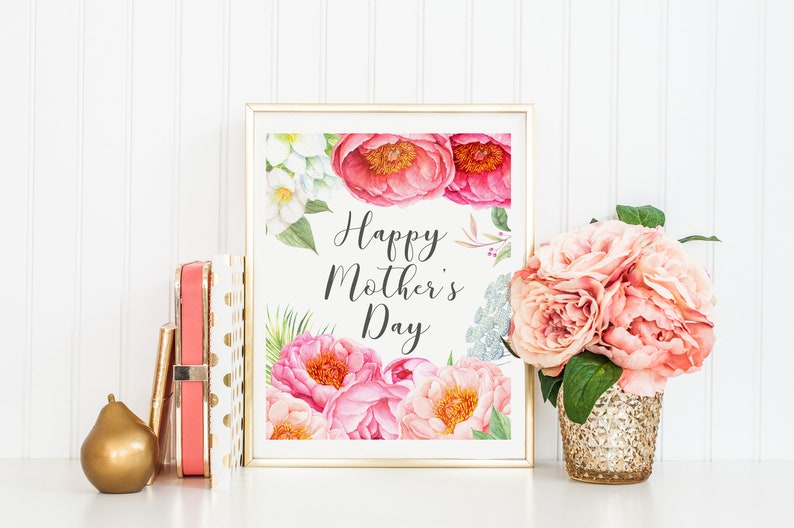 May include: A framed print with a watercolor illustration of pink and white flowers. The print says "Happy Mother's Day" in black script. The print is sitting on a white surface with a gold pear and a stack of pink and gold gift boxes.
