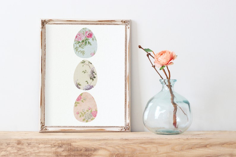 May include: A framed print of three pastel-colored Easter eggs with floral patterns. The eggs are arranged vertically in a row. The frame is white with a distressed finish.