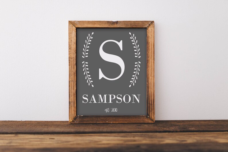 May include: Wooden framed sign with a gray background featuring a white letter "S" and the name "SAMPSON" below it. The sign also includes the text "est. 2010". Decorative white leaf designs frame the letter. The sign is displayed on a wooden surface.