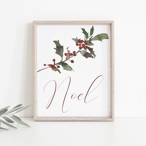 May include: A framed watercolor print featuring a sprig of holly with red berries and green leaves. The word "Noel" is written in a burgundy script font below the holly. The frame is a light wood color.