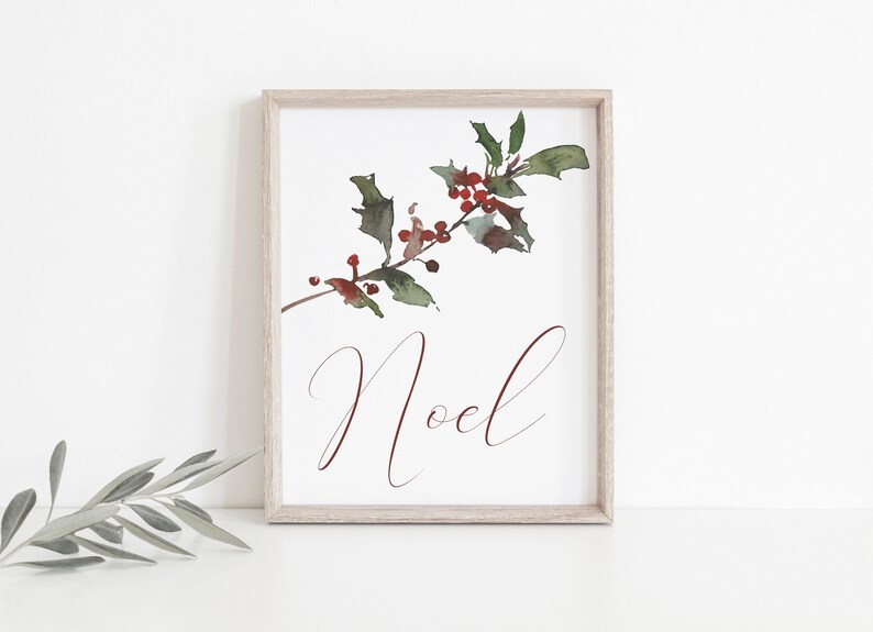 May include: A framed watercolor print featuring a sprig of holly with red berries and green leaves. The word "Noel" is written in a burgundy script font below the holly. The frame is a light wood color.