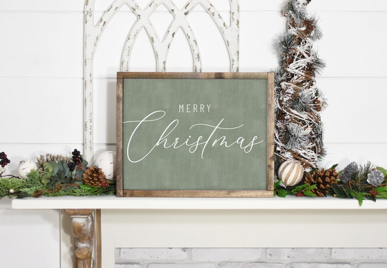 Pode incluir: Uma placa de madeira verde com o texto "Merry Christmas" em escrita cursiva branca. A placa est&aacute; sentada em uma lareira branca com uma pequena &aacute;rvore de pinho nevada e outras decora&ccedil;&otilde;es de Natal.
