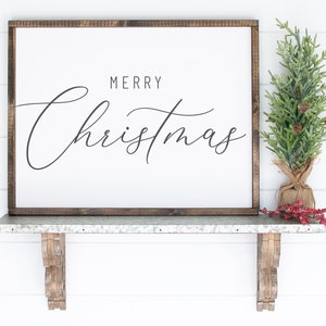 Pode incluir: Uma placa de madeira branca com moldura marrom, apresentando o texto "Merry Christmas" em escrita cursiva preta. A placa &eacute; exibida em uma prateleira de metal com suportes de madeira decorativos, e uma pequena &aacute;rvore de pinho artificial com bagas vermelhas &eacute; colocada na prateleira.