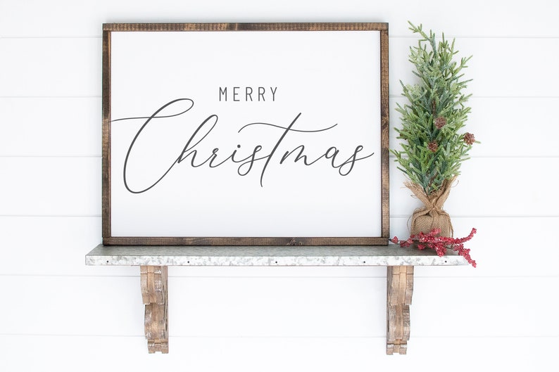 Pode incluir: Uma placa de madeira branca com moldura marrom, apresentando o texto "Merry Christmas" em escrita cursiva preta. A placa &eacute; exibida em uma prateleira de metal com suportes de madeira decorativos, e uma pequena &aacute;rvore de pinho artificial com bagas vermelhas &eacute; colocada na prateleira.
