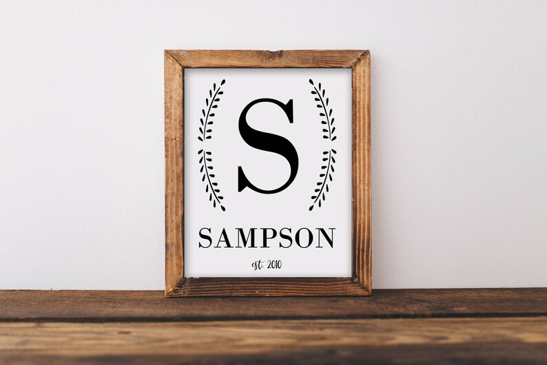 May include: A framed wooden sign with a white background. The sign features a large black letter "S" in the center, with the name "SAMPSON" below it. The text "est. 2010" is below the name. Decorative leaf sprigs flank the letter.