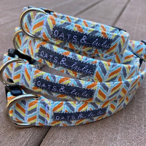 May include: Four dog collars in different sizes, stacked on top of each other. The collars are made of a light blue fabric with a chevron pattern in orange, yellow, and gray. Each collar has a black label with the text "Oats & Lulu" in white.