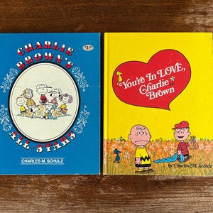 May include: Two vintage children's books featuring Charlie Brown and the Peanuts gang. One book has a blue cover with the title "Charlie Brown's All-Stars". The other book has a yellow cover with a red heart and the title "You're In Love, Charlie Brown".