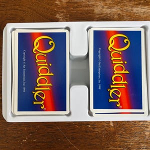 May include: Two decks of Quiddler cards in a white plastic tray. The card backs are blue with a yellow and orange logo. The text "Quiddler" is prominently displayed. Copyright Set Enterprises, Inc. 1998 is also visible.