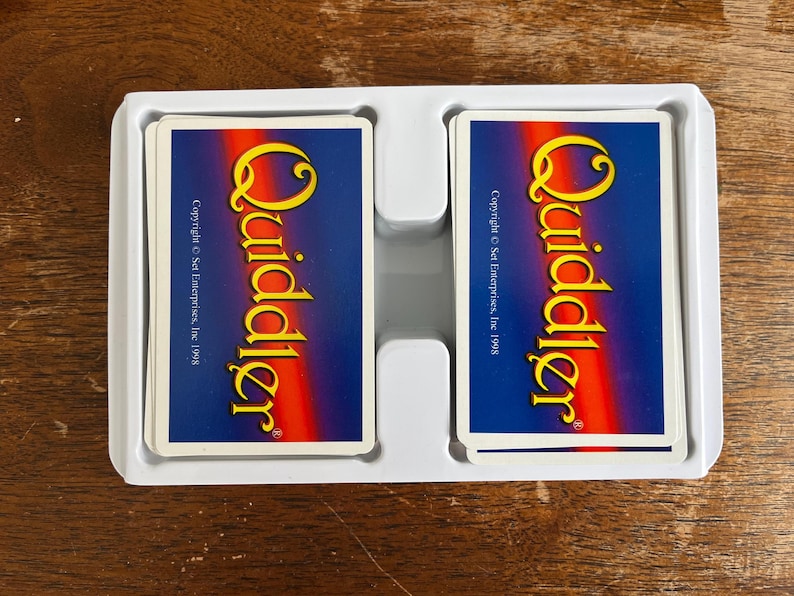 May include: Two decks of Quiddler cards in a white plastic tray. The card backs are blue with a yellow and orange logo. The text "Quiddler" is prominently displayed. Copyright Set Enterprises, Inc. 1998 is also visible.