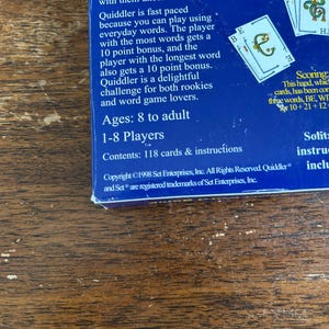 May include: A section of a blue game box with the word "Quiddler" visible, along with game instructions and card details. The box is on a wooden surface. The game is for 1-8 players, ages 8 to adult.