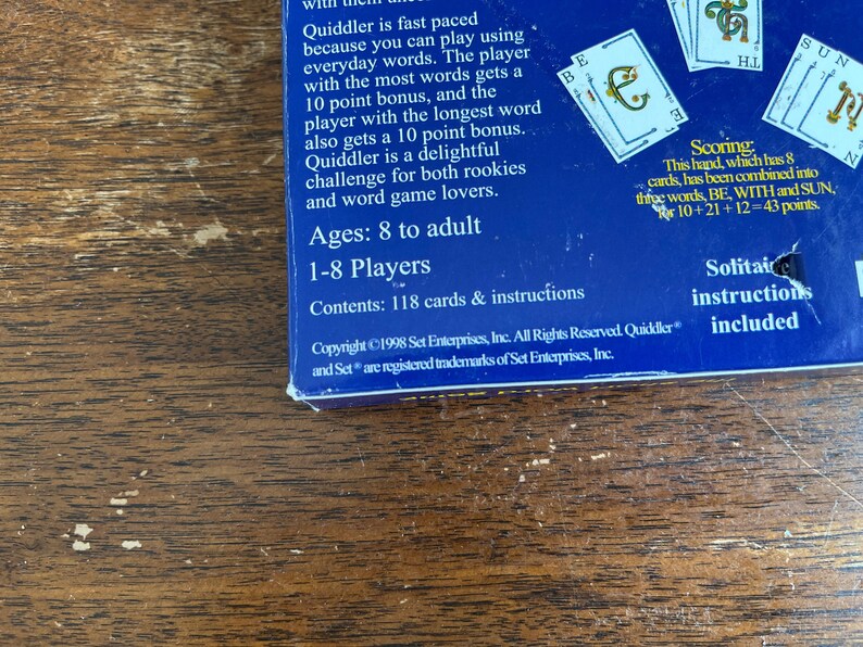 May include: A section of a blue game box with the word "Quiddler" visible, along with game instructions and card details. The box is on a wooden surface. The game is for 1-8 players, ages 8 to adult.