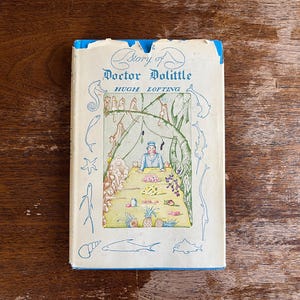 May include: Vintage hardcover book titled "Story of Doctor Dolittle" by Hugh Lofting. The cover features an illustration of a man at a table with animals and food. The book has a cream-colored cover with blue accents and illustrations.