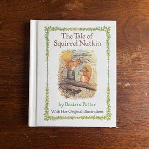 以下が含まれることがあります： ビクトリア・ポター著「リスのナトキンのお話」というタイトルのハードカバー本。表紙には2匹のリスのイラストが描かれています。本は白い表紙に緑色の花の縁取りがあり、木製の表面に置かれています。