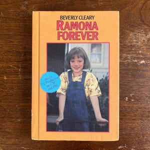 May include: A vintage copy of "Ramona Forever" by Beverly Cleary. The book has a light orange cover with the title in red and pink. The cover features a photo of a young girl in denim overalls. A blue sticker indicates it was a TV production.