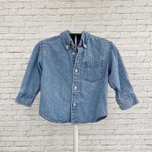 Könnte beinhalten: Ein hellblaues Jeanshemd mit Knopfleiste, langen Ärmeln mit geknöpften Manschetten, einem spitzen Kragen und einer einzelnen Brusttasche auf der linken Seite. Das Hemd hat eine Knopfleiste vorne mit weißen Knöpfen und einen abgerundeten Saum.