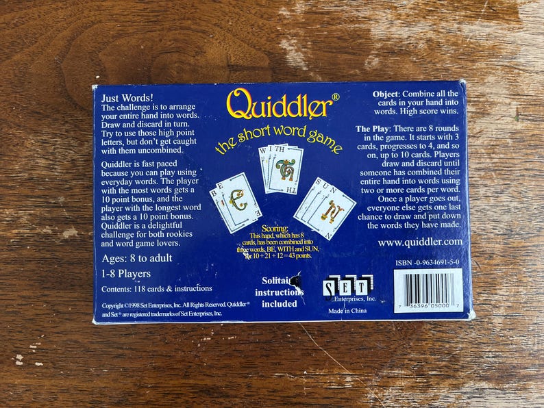 May include: The back of a Quiddler card game box, featuring the game's logo and instructions. The box is dark blue with white text and illustrations of cards. The game is for 1-8 players, ages 8 to adult.