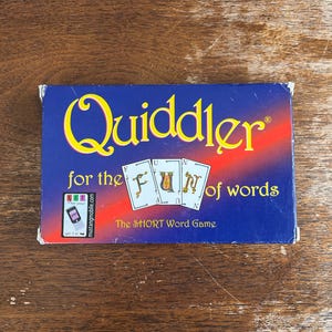 May include: A blue game box for "Quiddler" with yellow and red text. The box features the game's name, the phrase "for the FUN of words," and the tagline "The SHORT Word Game." Three playing cards are also visible.